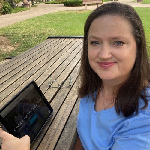 A woman sitting at a wooden table in a park, using a tablet with glasses placed beside it.