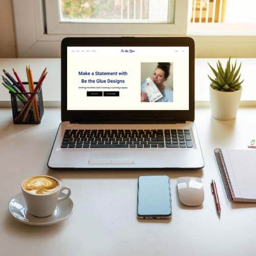 A neatly organized desk with a laptop open to a design advertisement, surrounded by a cup of coffee, a smartphone, notebook and plant, in front of a window.